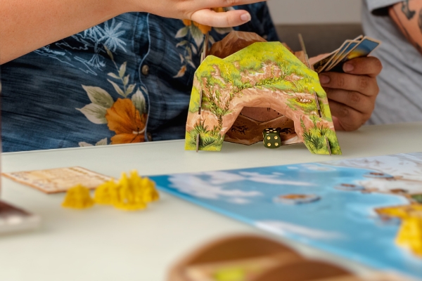 Players using the CATAN Dawn of Humankind promo dice tower, showing a cave‑style design with dice rolling out.