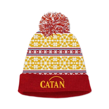 A red beanie with a yellow, blue, and white snowflake pattern and a red and white pompom. The beanie has the "Catan" logo on the bottom cuff.