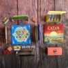 Catan Zip Edition components arranged on a wood surface, including the compact game board, resource trays, cards, dice, and the red and yellow carry bag.